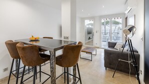 Uma sala de jantar moderna com uma mesa e quatro cadeiras, um sofá e um tripé em um espaço claro com paredes brancas e janelas amplas.