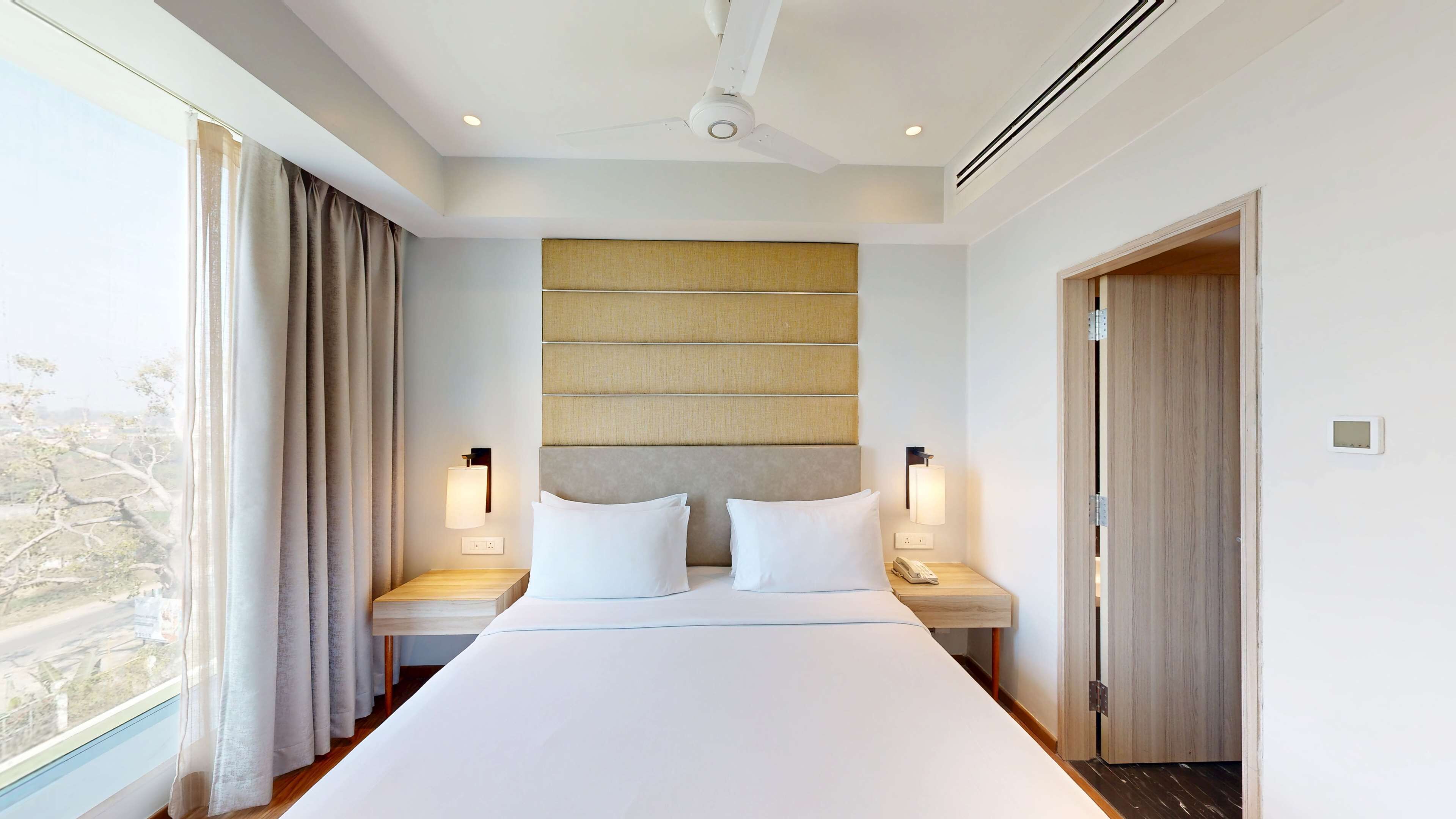 Habitación de hotel con una cama grande, dos mesitas de noche, cabecero y ventilador de techo, con vista al exterior.
