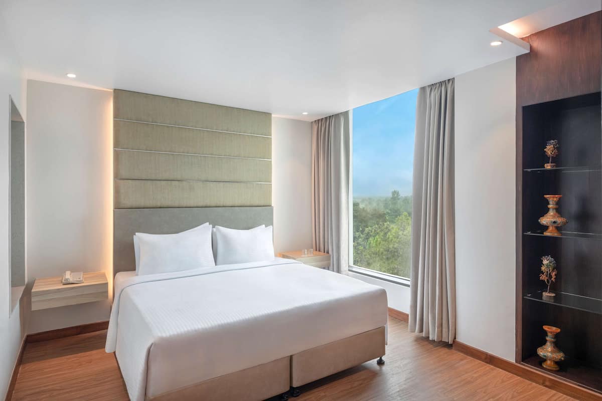 A modern hotel room with a large bed, a bedside table, a nightstand, a window with curtains, and a wall-mounted shelf with decorative items.