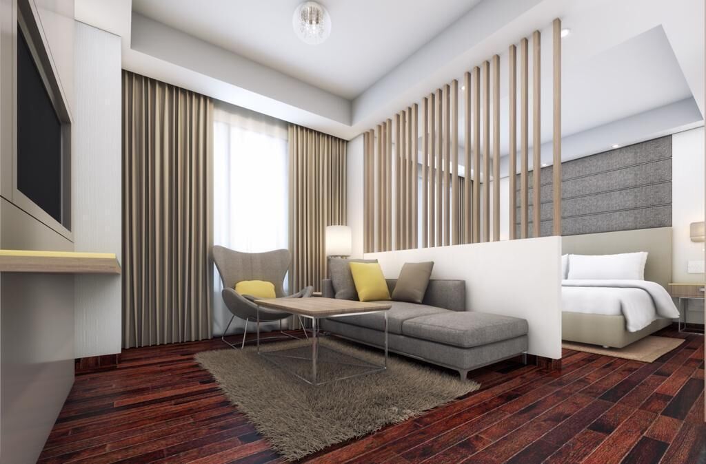 A modern hotel room with a grey sofa, a small table, a chair, a bed, and a large window with curtains.