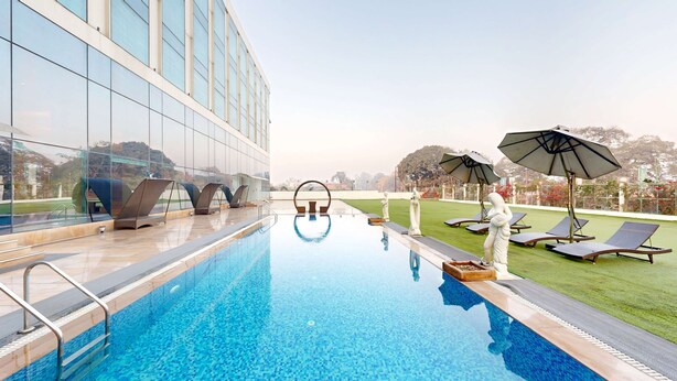 A swimming pool with lounge chairs and umbrellas, surrounded by a building with large windows. - Radisson Hotel Bareilly Airport (Bareilly)