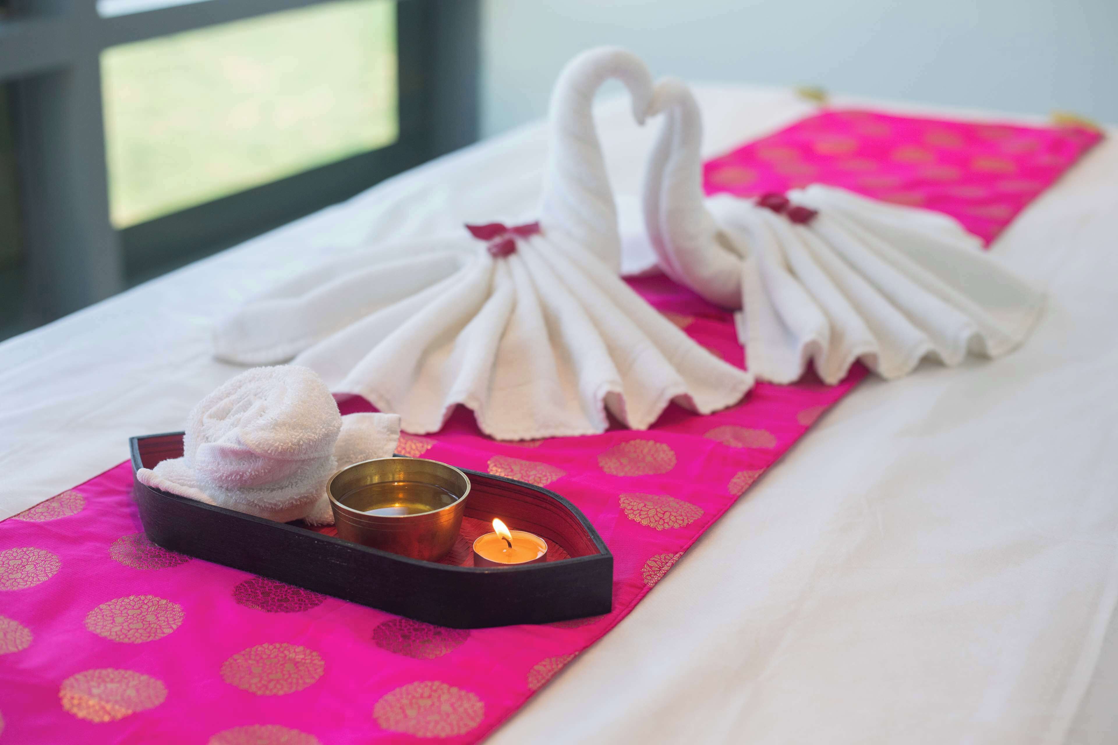 Un lit avec du linge de lit blanc, une couvre-lit à pois roses et un plateau avec une bougie allumée et des serviettes pliées.