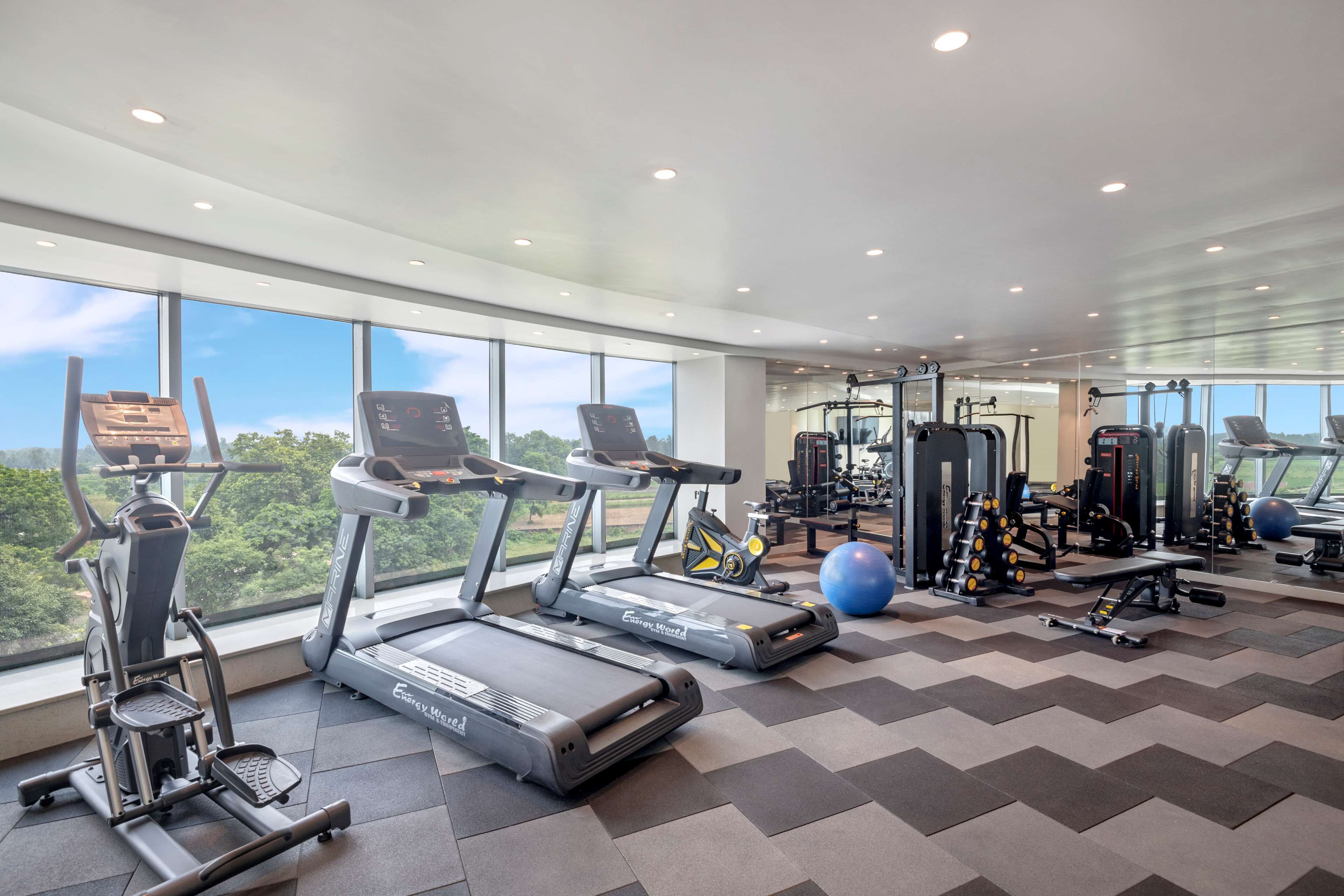 A gym with treadmills, elliptical machines, and weightlifting equipment, large windows, and a chevron-patterned floor.