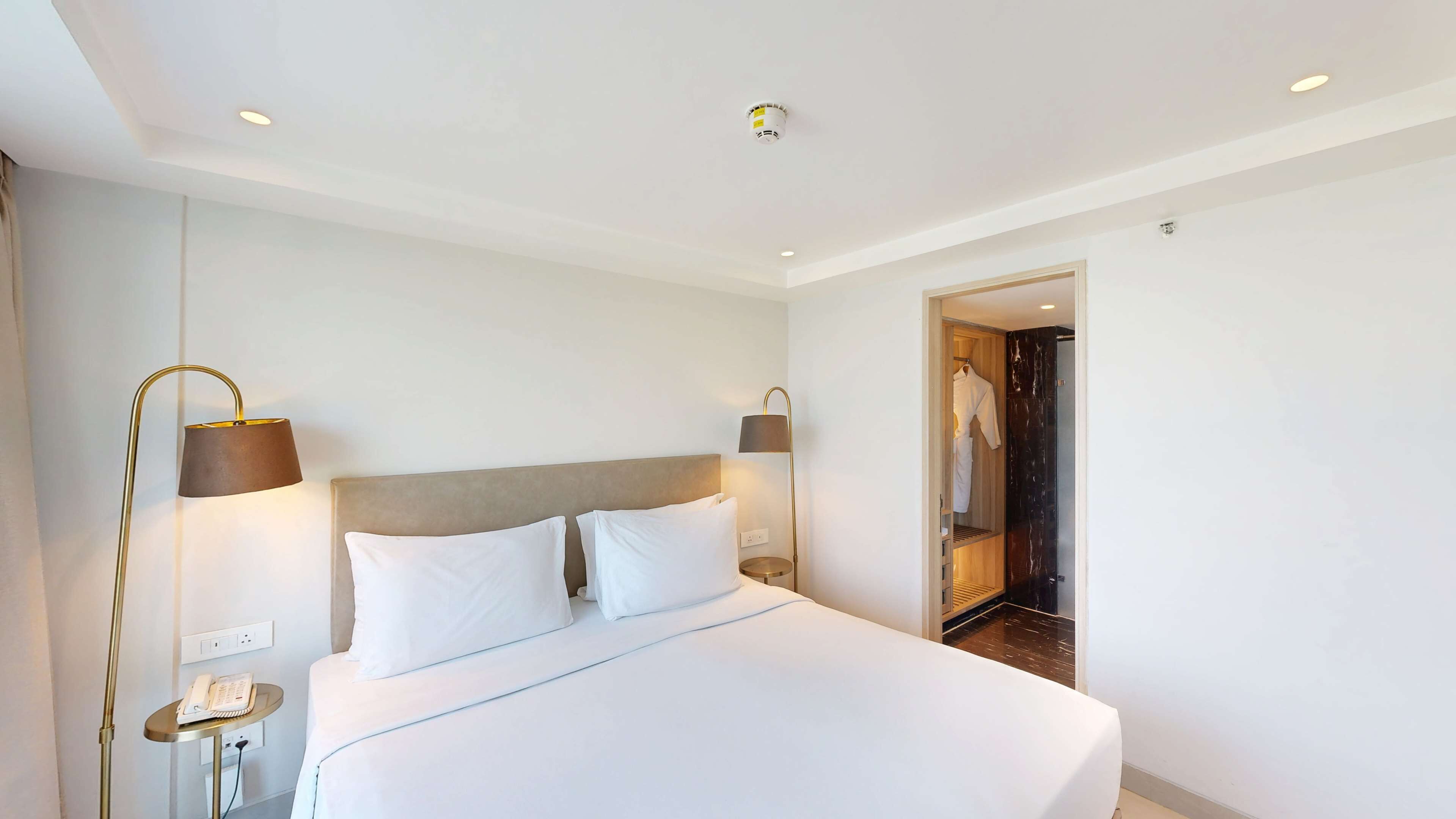 Habitación de hotel con una cama grande, dos lámparas de noche, un teléfono sobre una mesita y un armario vestidor con bata.