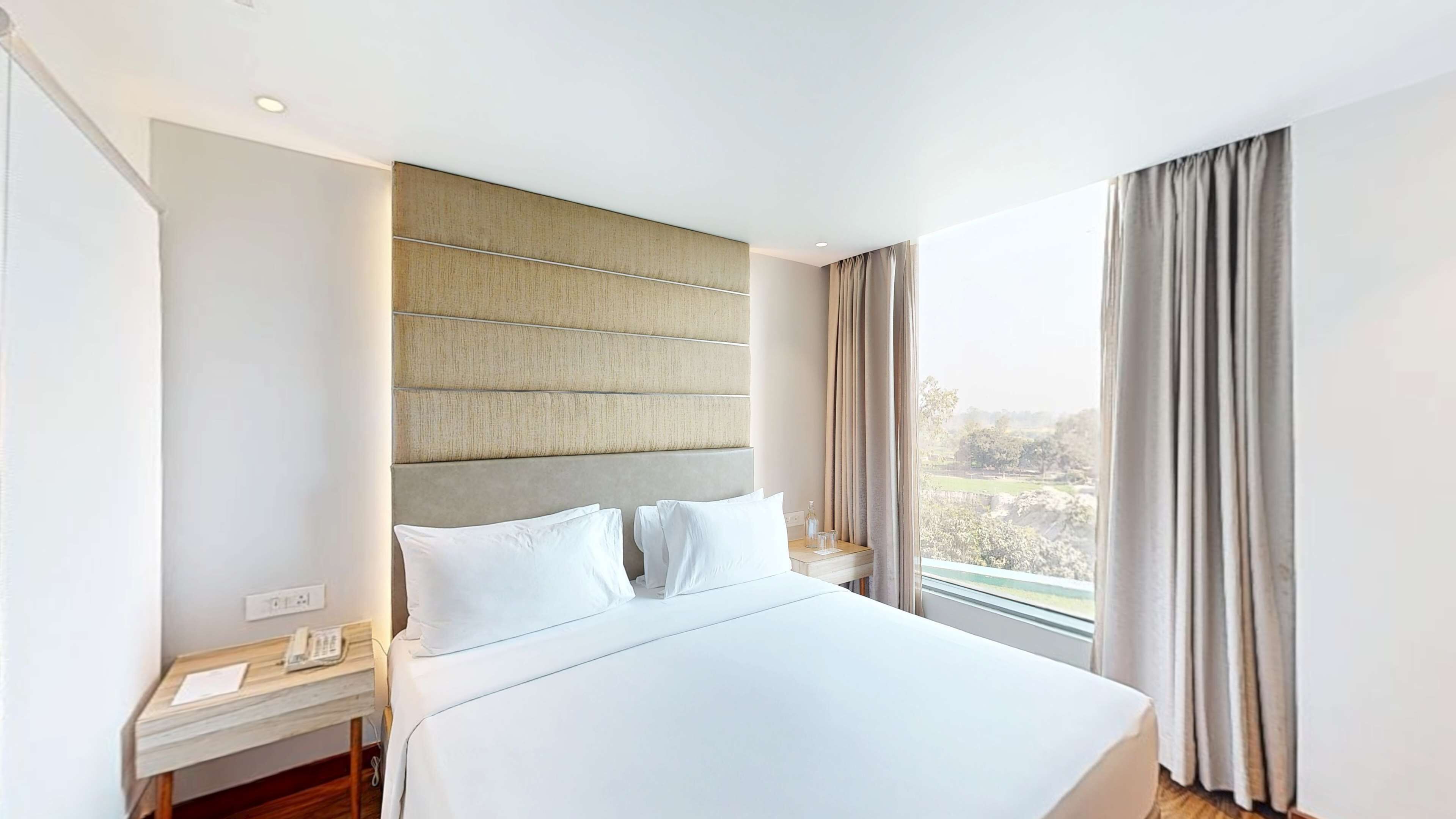A hotel room with a large bed, a bedside table with a phone, and a view of a landscape through the window.
