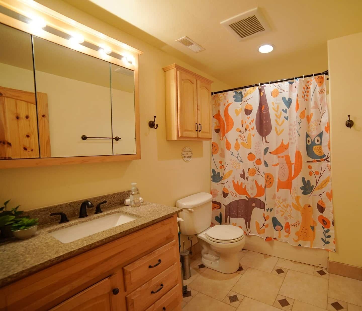 Un baño con tocador de madera, lavamanos, inodoro y cortina de ducha con diseño de motivos naturales.