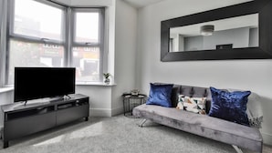 Un salon moderne avec un canapé gris, un téléviseur à écran plat posé sur un meuble TV noir, une petite table ronde avec une lampe et un grand miroir accroché au mur.