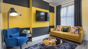 A modern living room with a blue armchair, a yellow sofa with colorful cushions, a mounted TV, and a black lamp. - ★Heart of the City★Totally Upgraded ✔ Sleeps 10 (Sheffield)