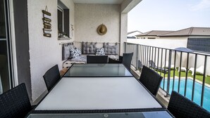 Een balkon met een tafel met glazen blad, rieten stoelen, een gewatteerd bankje en een zwembad.