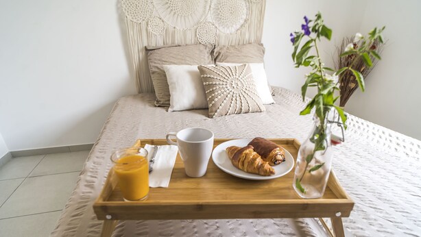 Een net opgemaakt bed met een decoratief hoofdbord, een dienblad met een croissant, een mok en een glas sinaasappelsap op het bed.