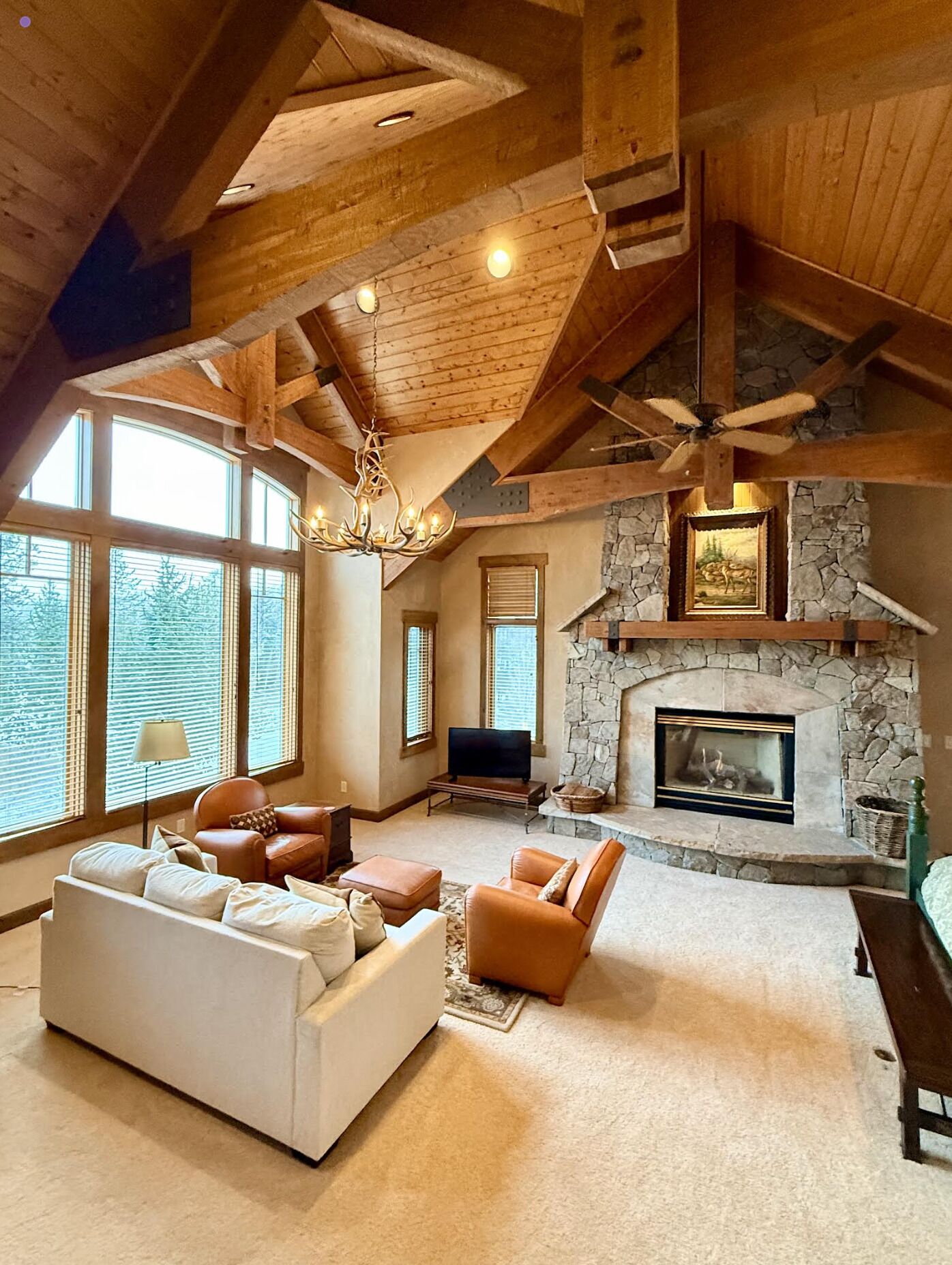 A spacious living room with a stone fireplace, large windows, and a high ceiling with exposed beams.