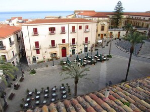 Una plaza con mesas al aire libre, un edificio con contraventanas rojas y una palmera.