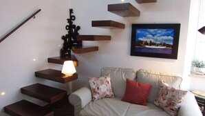 A modern living room with a beige sofa, a staircase with dark wooden steps, and a wall-mounted picture of a coastal scene. - Arias la Suite del Duomo - Sea View Terrace (Cefalù)