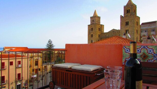 A balcony with a wooden bench, a wine bottle, and a glass. In the background, there is a building with two towers and a tiled roof. - Arias la Suite del Duomo - Sea View Terrace (Cefalù)
