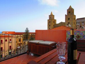 A balcony with a wooden bench, a wine bottle, and a glass. In the background, there is a building with two towers and a tiled roof. - Arias la Suite del Duomo - Sea View Terrace (Cefalù)