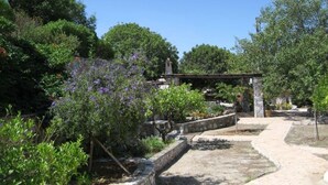 Een pad van stenen leidt naar een gebouw met stenen en een dak, omringd door weelderige begroeiing en bloeiende planten.