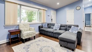 Un salon avec un canapé d’angle gris, un fauteuil à motifs, une petite table d’appoint en bois et une grande fenêtre avec des rideaux.