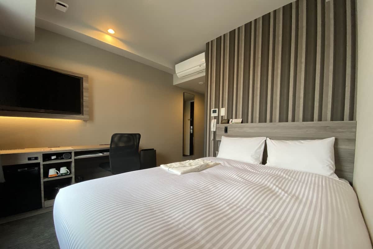 A hotel room with a bed, a desk, a chair, a TV, and an air conditioning unit.