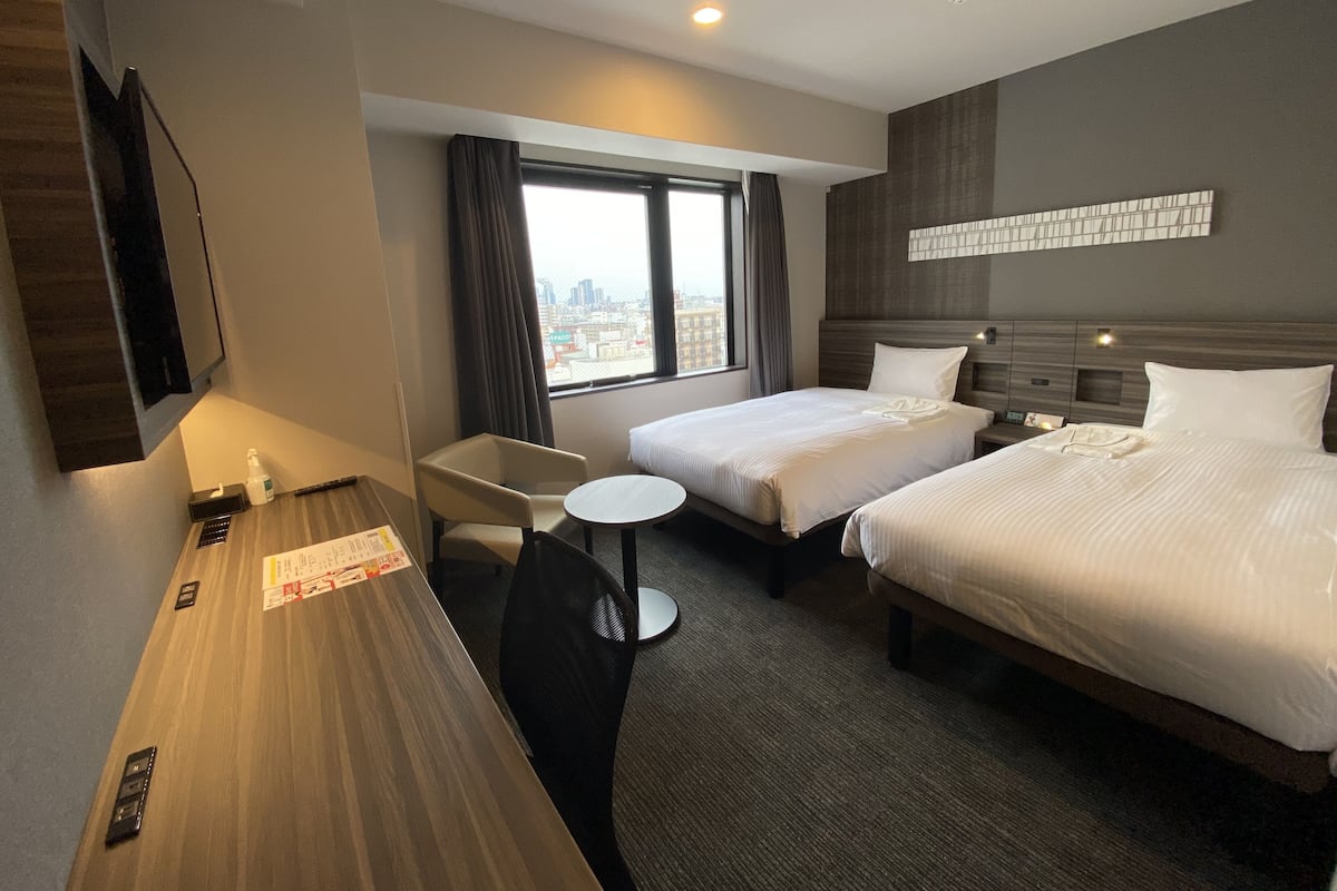 A hotel room with two beds, a desk, a chair, and a window with a city view.