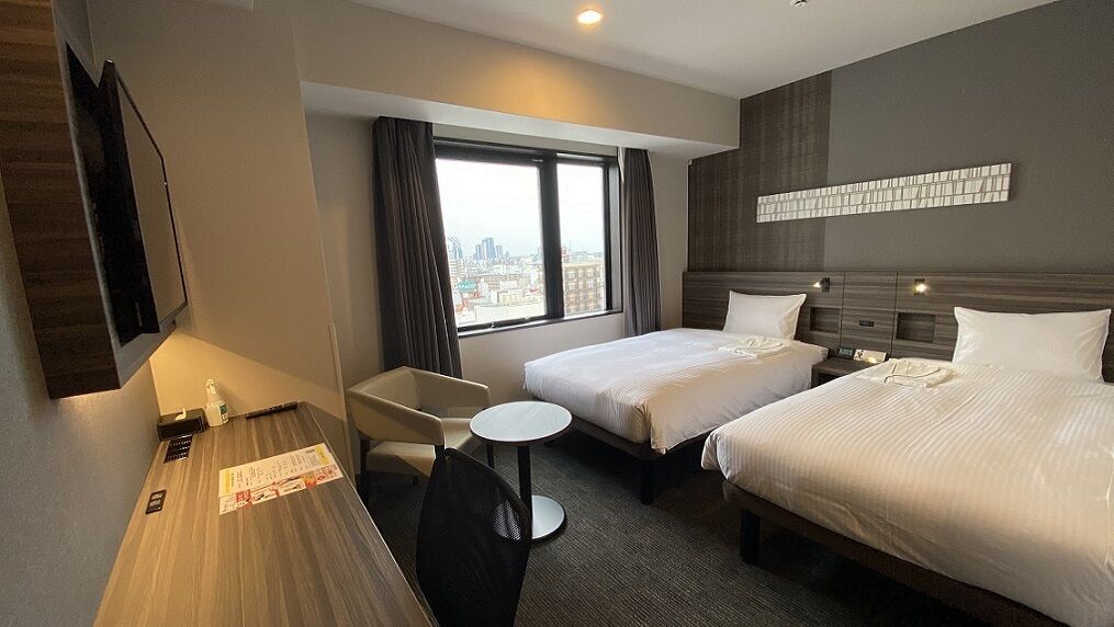 A hotel room with two beds, a desk, a chair, a small table, and a window with a city view.