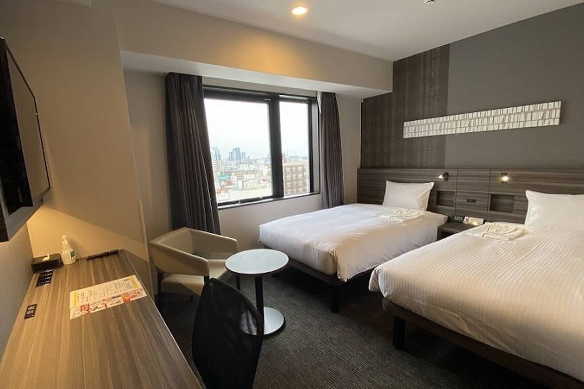 A hotel room with two beds, a desk, a chair, a small table, and a window with a city view.