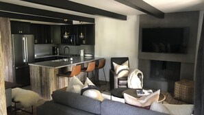 Uma sala de estar moderna com um sofá cinza, uma mesa de centro e uma lareira. Uma cozinha com armários escuros e eletrodomésticos de aço inoxidável é visível ao fundo.