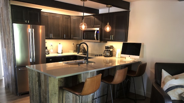 Cozinha moderna com armários escuros, eletrodomésticos de aço inoxidável, ilha central com banquetas e iluminação pendente.