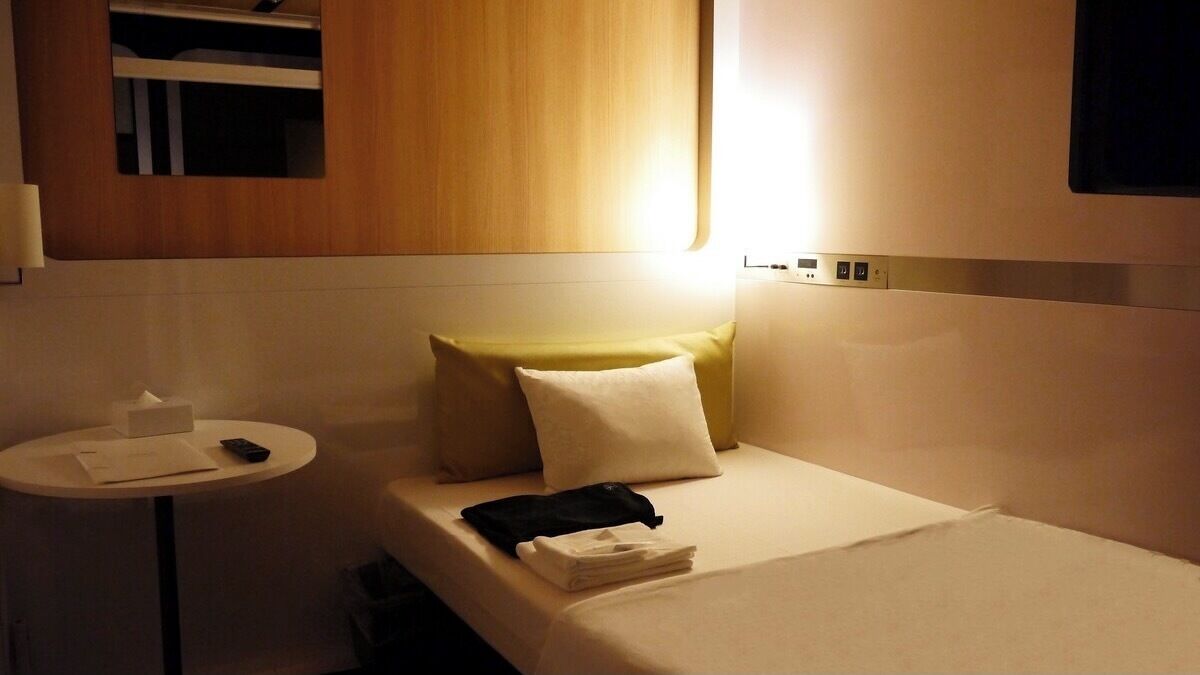 A hotel room with a bed, a bedside table with a remote control, and a small table with a tissue box and papers.