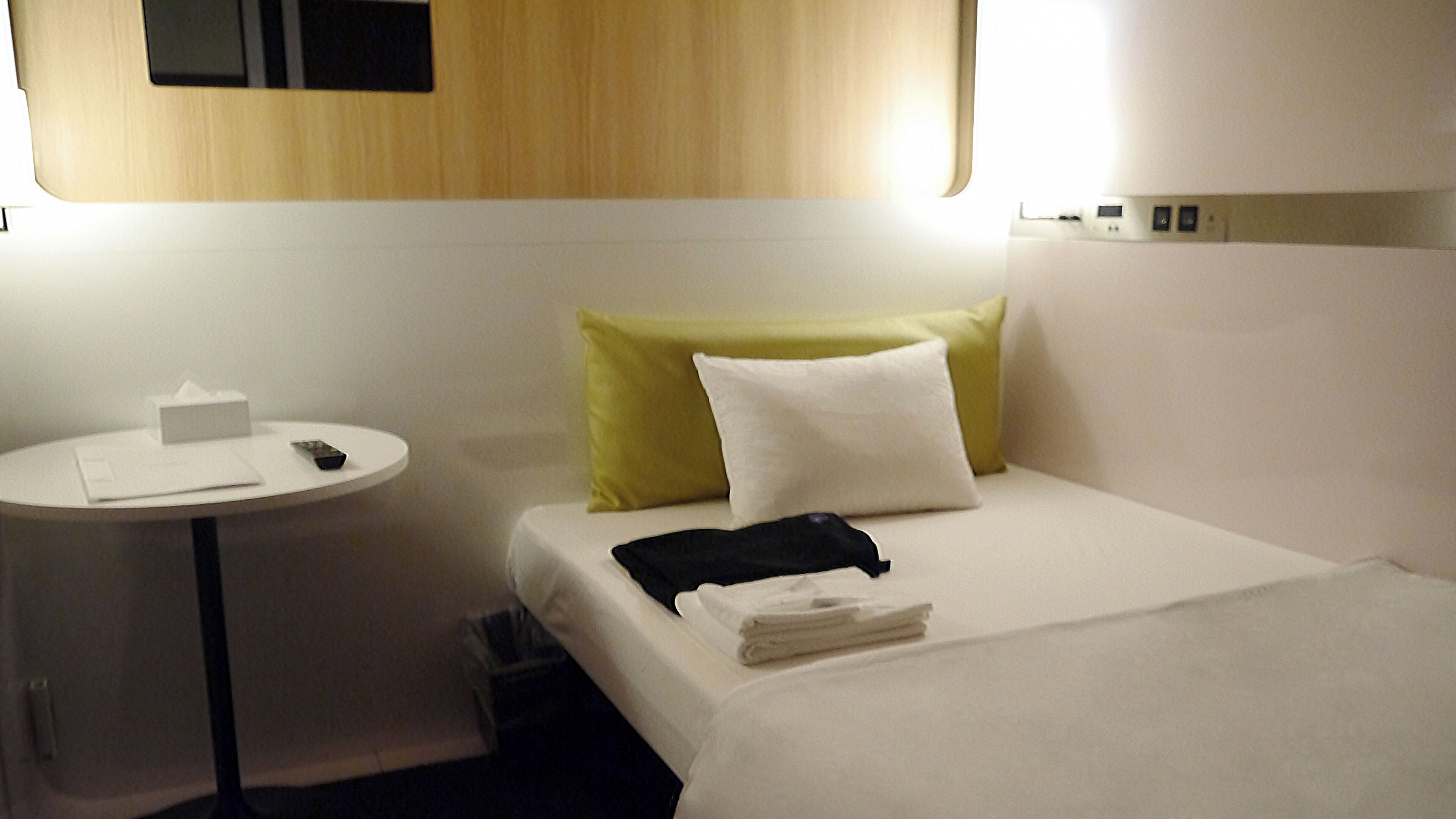 A hotel room with a bed, a bedside table, a small table, and a wall-mounted control panel.