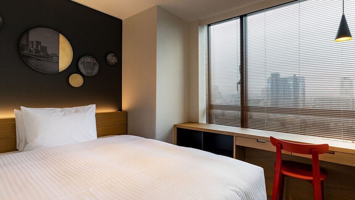 Une chambre d’hôtel avec un lit, un bureau, une chaise et une fenêtre avec des stores.