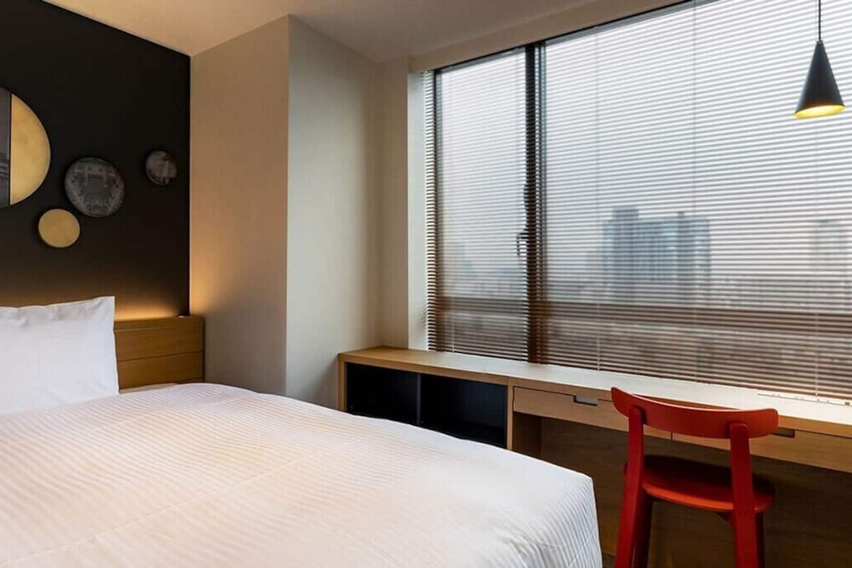 A hotel room with a bed, a desk, a chair, and a window with blinds.