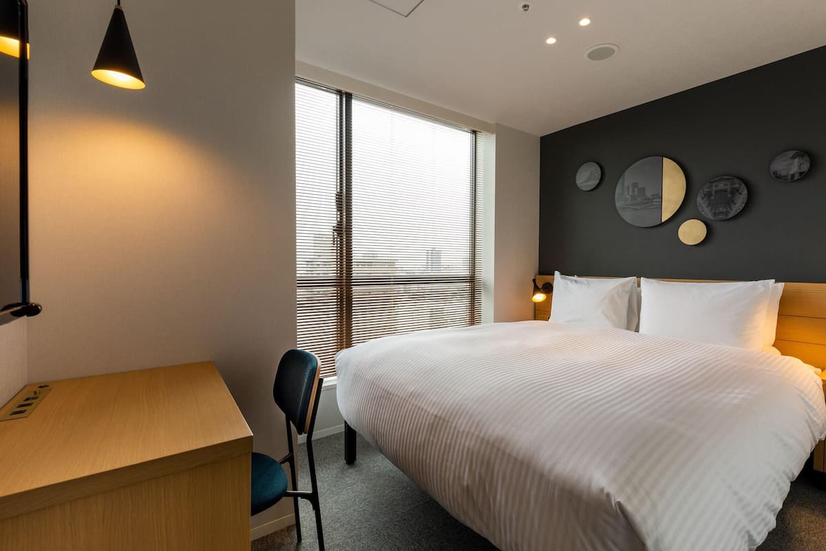 A modern hotel room with a large bed, a desk, a chair, and a window with blinds.