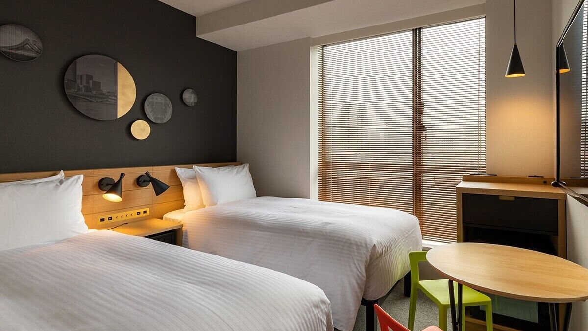 Une chambre d’hôtel avec deux lits, une petite table, une chaise, un bureau et une grande fenêtre avec des stores.