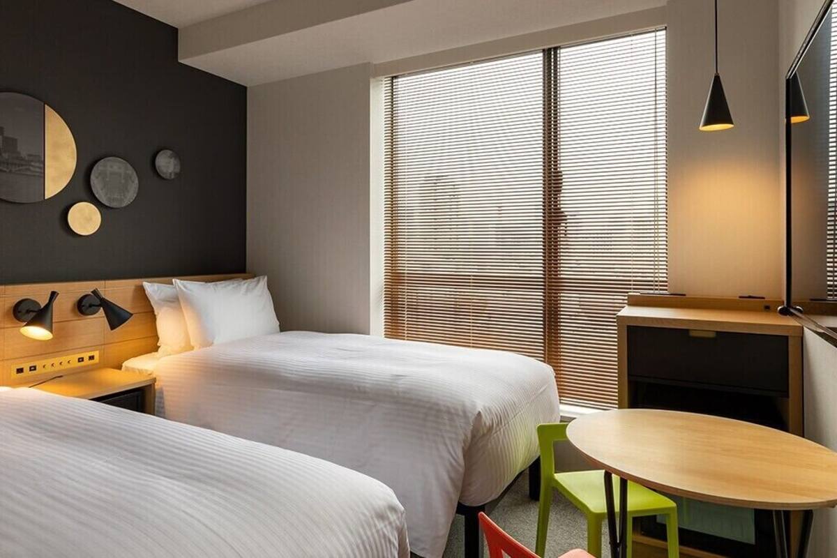 A hotel room with two beds, a small table, a chair, a desk, and a large window with blinds.