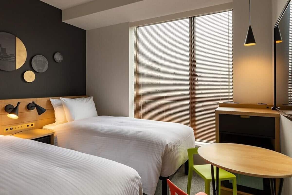 Quarto de hotel com duas camas, uma pequena mesa, uma cadeira, escrivaninha e uma janela grande com persianas.