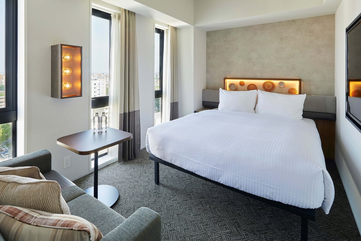 A hotel room with a large bed, a small table, a sofa, and a view of the city through the window.