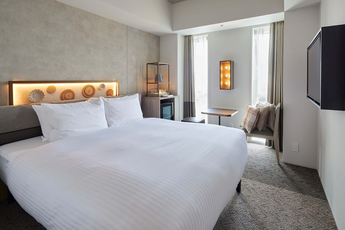 A modern hotel room with a large bed, a desk, a TV, and a window with curtains.