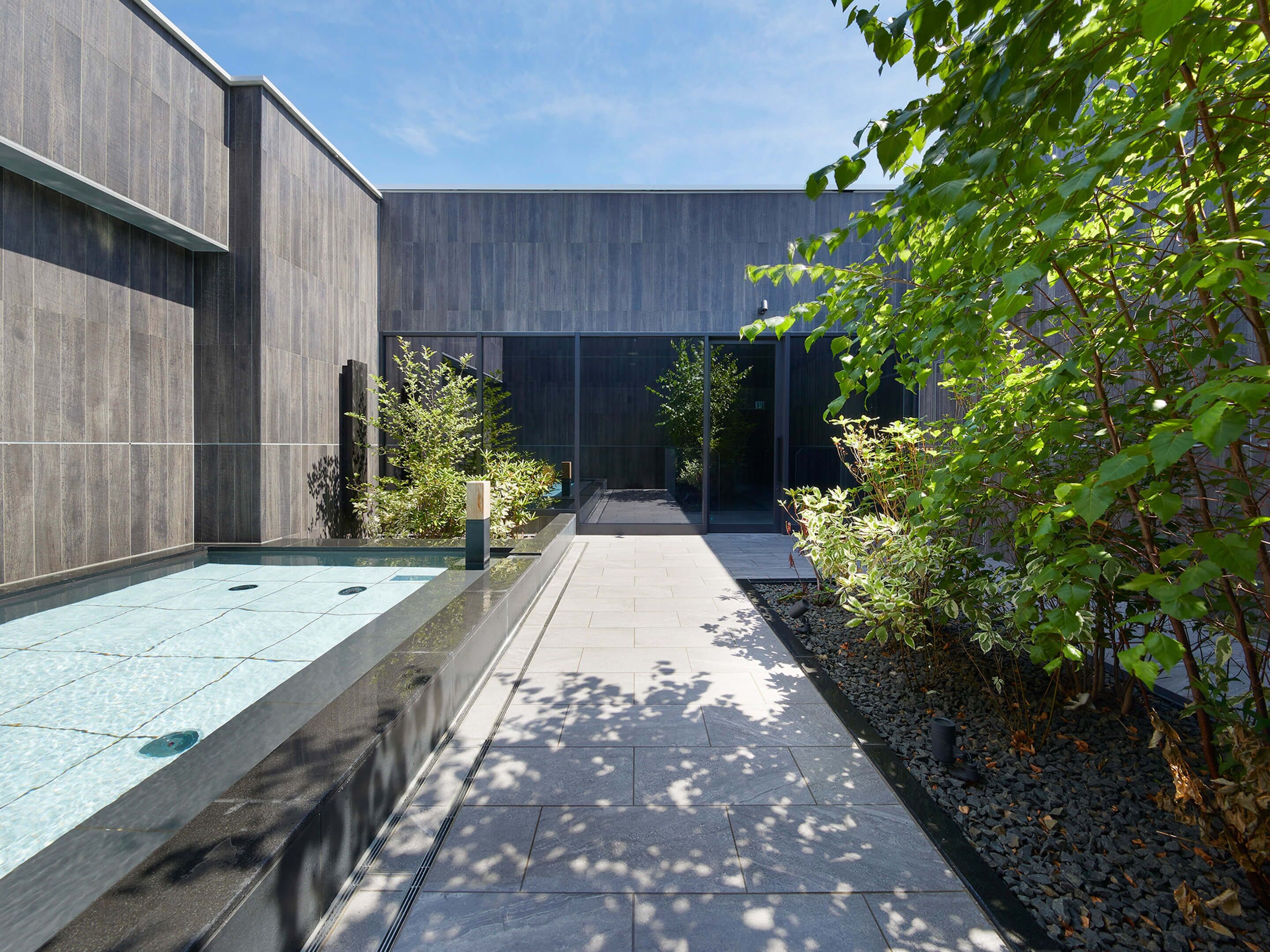 A modern outdoor pool area with a clear pathway, surrounded by greenery and a building with large windows.