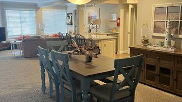 A dining area with a wooden table and chairs, a chandelier, and a kitchen in the background.
