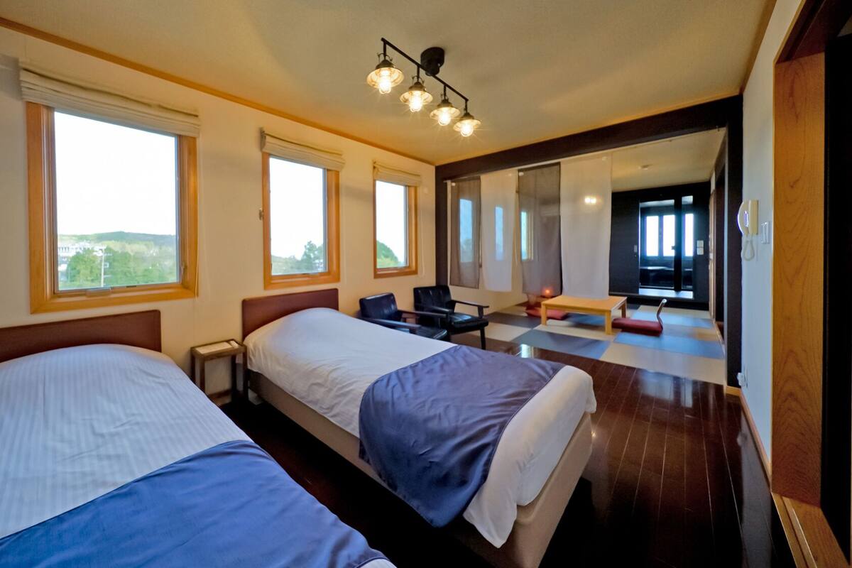 Ein Hotelzimmer mit zwei Betten, großen Fenstern, einem Schreibtisch und einem Sessel.