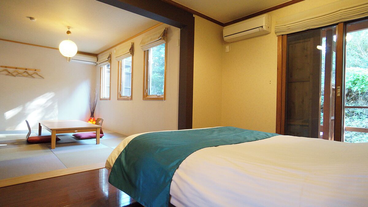 A bedroom with a bed, a small table, and a chair. There are windows and an air conditioning unit.