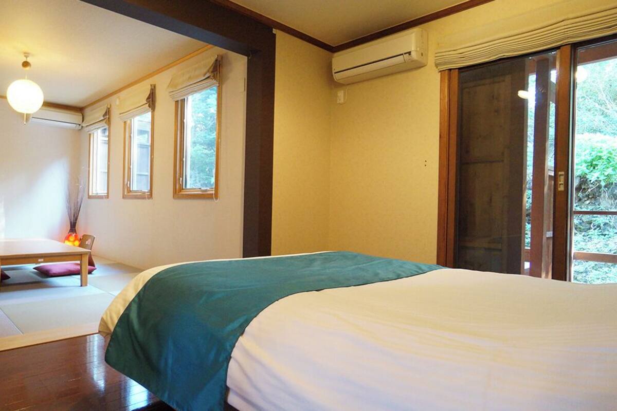 Ein Schlafzimmer mit einem Bett, einem kleinen Tisch und einem Sessel. Es gibt Fenster und eine Klimaanlage.