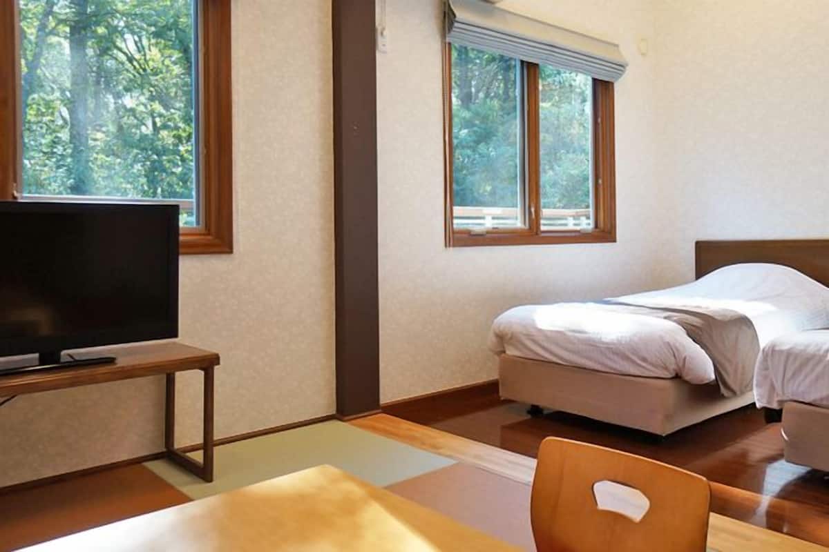 Ein Hotelzimmer mit zwei Betten, einem Fernseher auf einem Schrank und einem Fenster mit Blick auf Bäume.