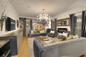 A modern living room with a grey sofa, a coffee table, and a TV. There is a chandelier, a wall with a clock mural, and a window with curtains.