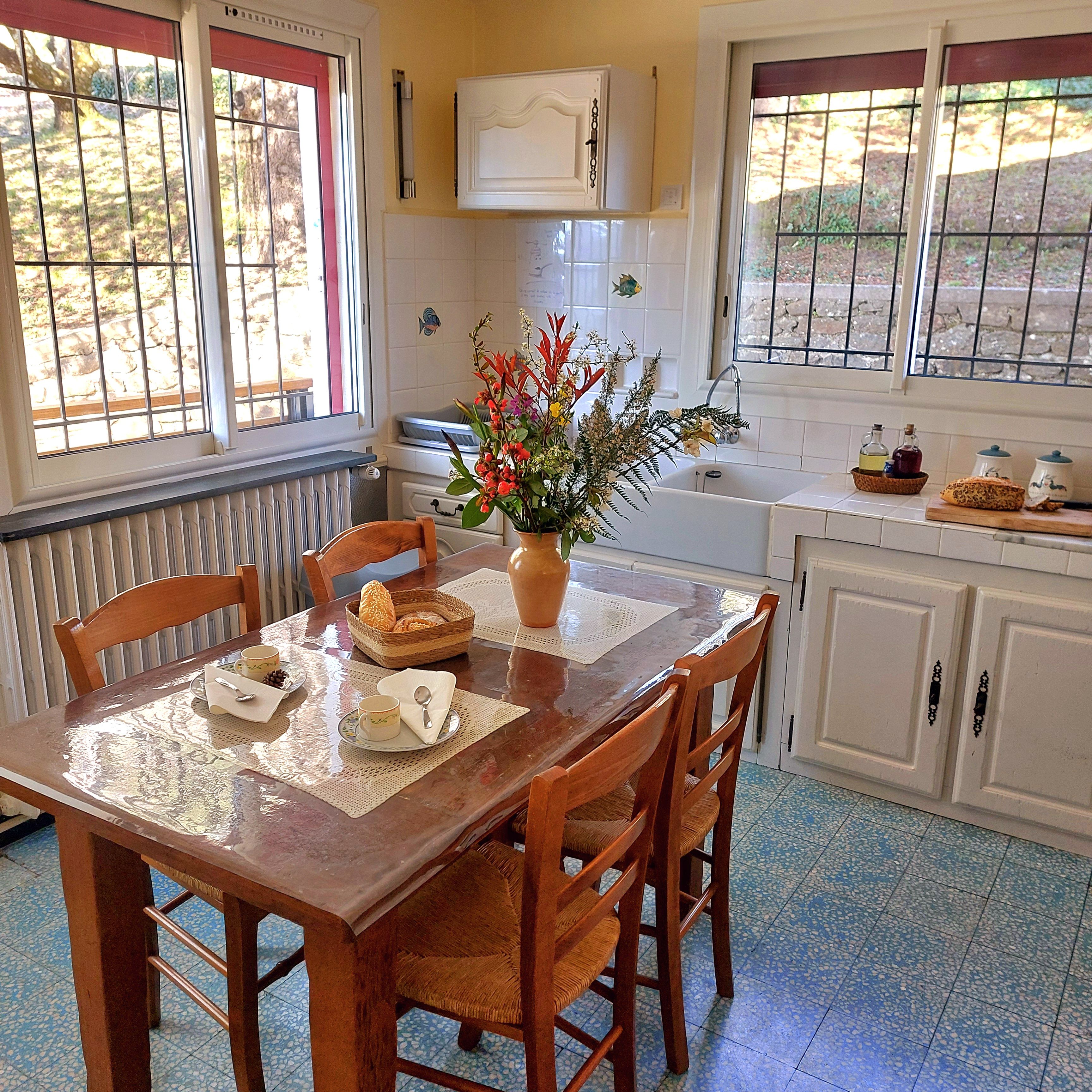 Un coin repas avec une table en bois dressée pour deux personnes, un vase de fleurs et une cuisine en arrière-plan.