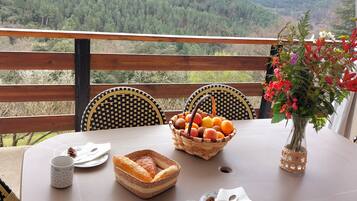 A table set with a basket of fruit, a vase with flowers, and a mug, overlooking a forested mountain range.