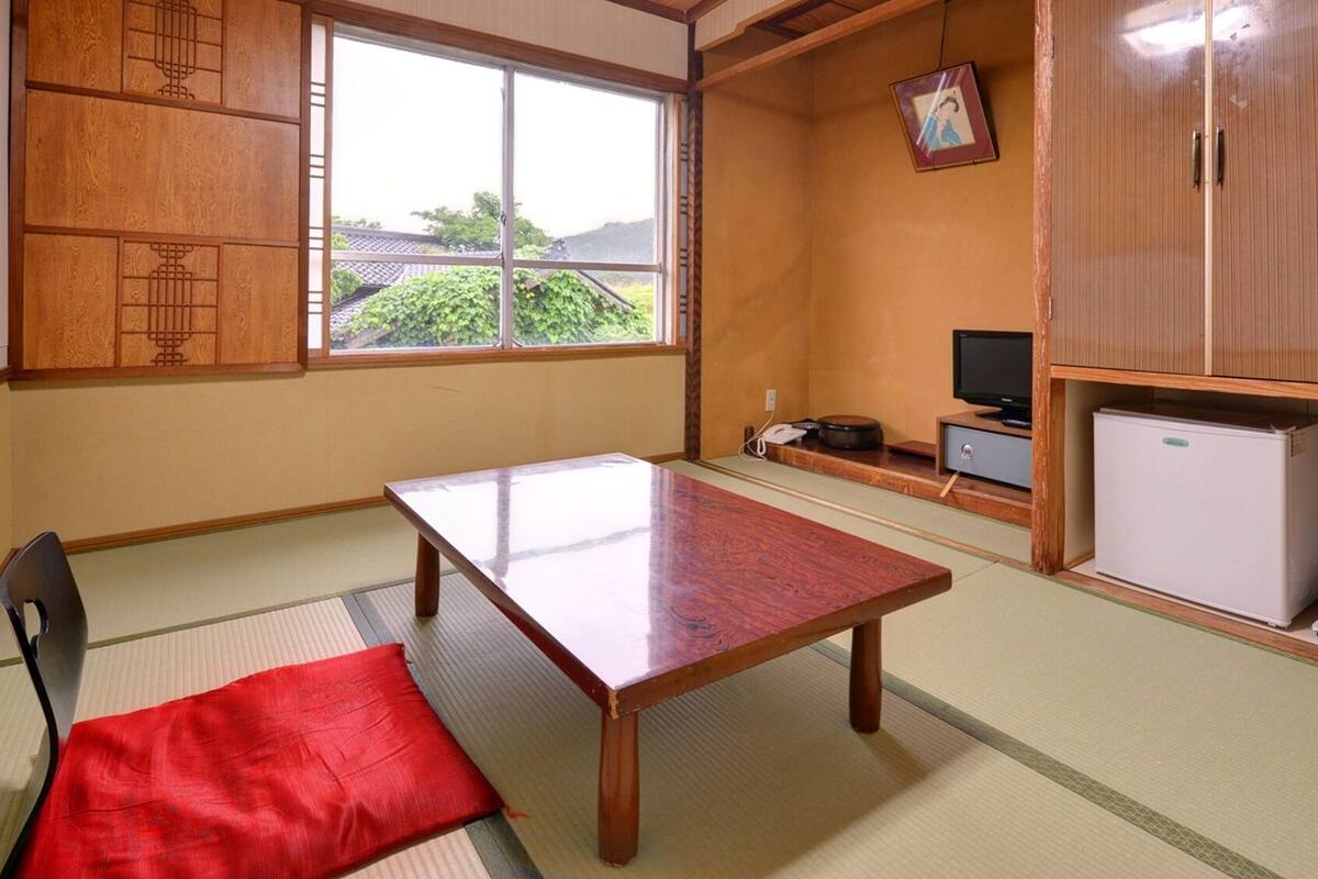 Una habitación japonesa tradicional con suelo de tatami, una mesa baja, una silla, un televisor y un refrigerador.