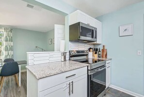 Een moderne keuken met witte kastjes, een granieten aanrechtblad, een magnetron, een oven en een eethoek met een stoel en tafel.