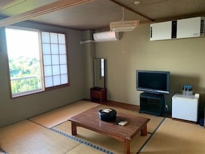Een kamer met tatami vloerbedekking, een televisie, een kleine koelkast en een raam met uitzicht op groen.