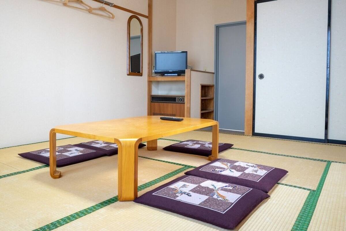 Una habitación con esteras de tatami, una mesa de madera, un televisor en un estante y una puerta.